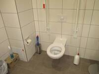 Eine weiße Toilette an einer weiß gekachelten Wand, auf beiden Seiten ist ein Haltegriff.