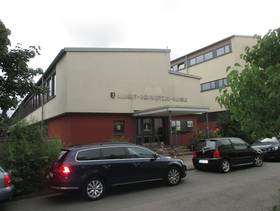 zwei bei einander stehende Gebäude, beim linken Gebäude ist der Schriftzug "Albert Schweitzer Schule " über dem überdachten Eingang, davor ist eine Straße mit parkenden Autos
