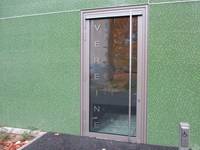 einflügelige Glastür mit senkrechten Griffstange. Die Tür ist in eine grüne, kleingeflieste Wand eingelassen
