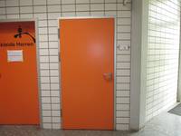 orangefarbenene Tür in einer weiß gekachelten Wand, links neben der Türe eine weitere orangene Tür, mit dem Schriftzug Umkleide Herren