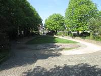Im Vordergrund ist ein Zugangsweg zu einer Parkfläche mit Rasen und einem Brunnen vor dem Rasen, der Rasen ist von einem Weg umgeben. Rechts und links an den Rändern des Parkes stehen mehrere Bäume die einen Schatten auf die Innenfläche werfen