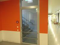 Eine Glastür in orangefarbenen Wand mit weißem Sockel, rechts davon langer Flur