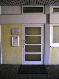 weiße mit Glaselementen versehen Tür an einer gelblichen Wand, links Praxisschild und Briefkasten, rechts Fensterausschnitt, im Vordergrund Natursteinplatten und Bodenmatte