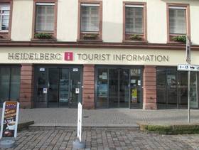 mehrstöckiges Gebäude mit einer Ladefront mit zwei Glasschiebtüren. Darüber Schriftzug "Heidelberg Tourist Information". Vor der Tür großes Podest als Abgrenzung zum Platz