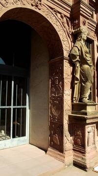 Reich verzierter Sandsteinbogen, rechts daneben steht eine in die Fassade integrierte Statue auf einem Sockel. Am Torbogen beginnt das Podest (als einzelne Stufe beschrieben). Die Tür mit Metallstreben ist in der Fassade zurückversetzt
