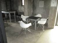 Hoher Raum mit Boden und Wänden aus Beton. In der Bildmitte steht ein kleinder quadtratischer Tisch mit 4 weißen Sesselstühlen. Links im Hintergrund  ein hoher Tisch mit einer hohen Sitzbank 