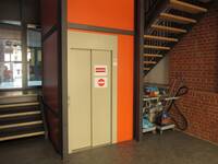 Ein heller Aufzug in einer orangefarbenen Wand. Links  ist eine aufwärts führende Treppe, rechts vom Aufzug die offene Rückseite einer weiteren aufwärts führenden Treppe, darunter stehen zwei Putzwägen