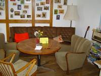 Auf dem Bild sieht man einen Teil des Aufenthaltsraum, mit einem Tisch mehreren Sesseln und einer Couch, an der Wand hinter der Couch sind etliche Bilder aufgehängt