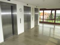Drei geschlossene Aufzüge mit Metalltüren nebeneinander. In der Mitte zwischen zwei Aufzügen hängt eine Uhr. 