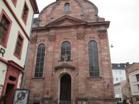 Aussenfront der Kirche aus Sandstein, in der Mitte im Erdgeschoss die Eingangstüre, rechts und links längliche Fenster und einige Verzierungen, über der Tür eine steinerne Statue