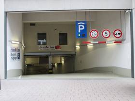 Offene Abfahrt in Tiefgarage. Rechts von Decke abgehängte schwarz-rotes Brett mit 3 Schildern: Einfahrtshöhe 2,1 m, 5 km/h, Anhänger verboten