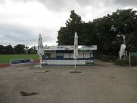 Auf einem Platz rechts vom Fußballfeld steht ein kleiner rehcteckiger Kiosk mit mehreren Verkaufsfenstern. Davor sind zwei geshclossenen Sonnenschirme. Rechts vom Kiosk sind Büsche und Bäume