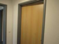 Tür in heller Holzoptik mit dunkelgrauem Rahmen in weißer Wand. Links daneben offen stehende baugleiche Tür zu Flur