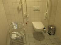 Ein weißes WC an einer weiß gekachelten Wand, mit Haltegriffen rechts und links. Neben der Toilette steht ein Stuhl aus Edelstahl mit Armlehnen.