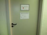 Eine helle Tür in einer weißen Wand. Auf der Tür sind zwei Schilder angebracht, eines mit der Aufschrift "Sitzungszimmer", das andere mit Raumbelegungsplan