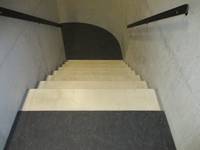 Nach unten führende Treppe aus hellem Stein, der Boden davor  und das anschließende Podest ist aus dunklem Stein, rechts und links ist ein Handlauf, das Podest ist abgerundet