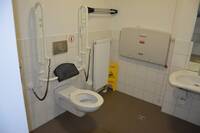 Weiße Toilette mit Rückenlehne an linker, weiß gekachelter Wand. HAltegriffe an beiden Seiten hochgeklappt. Rechts neben WC-Sitz schmaler Heizkörper. Rechts daneben hängt über Eck der ausklappbare Wickeltisch