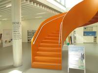Orangefarbene Wendeltreppe. Neben der Treppe ist eine Säule mit Hinweis, welche Räume sich auf welchen Ebenen befinden