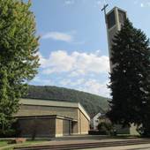 Ein Flachdachgebäude, etwas daneben ein Kirchturm mit einem Großen freistehenden Kreuz am oberen Ende, davor ein Platz und rechts und links ein Baum