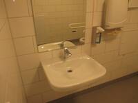 Weißes Waschbecken an weiß gekachelter Wand. Über dem Waschbecken hängt ein Spiegel. Rechts daneben hängt ein Spender für Flüssigseife oder Desinfektionsmittel, daneben ein Papierhandtuchspender 