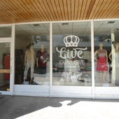 Links weiße Glastür, daneben dreigeteiltes, großes Fenster mit Schaufenster-Puppen. Mittig auf dem Fenster großes, weißes Logo mit Krone und Namenszug der Boutique.