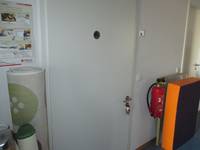 Weiße Tür in einer weißen Wand, auf der Tür ein rundes Schild. Rechts von der Tür eine zweifarbige Turnmatte. Dahinter ein roter Feuerlöscher. Links von der Tür eine zusammengerollte Matte und ein Plakat