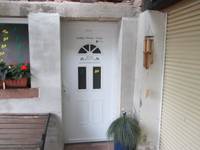 Weiße einflügelige Eingangstür, ein Stück daneben ein Fenster, unter dem Fenster steht eine Bank, rechts vor der Tür eine Pflanze in einem Kübel