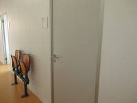 geschlossene hellgraue Tür in einer weißen Wand. Links davon ein Türschild, zwei Klappsstühle und eine offenstehende Tür. Der Boden ist aus orangenfarbenen Linoleum.