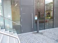 geschlossene Eingangstür mit senkrechter Griffstange, rechts davon eine Metallsäule, rechts und links Glasscheiben. Links vom Nebeneingang führen einige Stufen nach oben