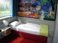 Teilansicht des Zimmers, eines der zwei Einzelbetten mit rotem Bettkasten. Links am Bett ist ein Nachtkästchen, darüber ist das Fenster, über dem Bett hängt ein großes, abstraktes, buntes Bild  