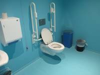 hoch gekachelter Raum mit blauen Wänden und Boden. Weißes Hänge-WC mit einem Haltegriff rechts und links. Links vom WC ist ein weißer Handtuchhalter, rechts stehen zwei Mülleimer an der Wand