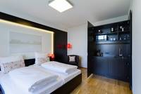 Raum mit Doppelbett in der Zimmermitte, über dem Bett ist ein Bild, daneben ein Sessel mit einer Stehlampe, im rechten Eck ist ein offenstehnder Schrank mit einer Küche darin