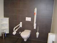 Auf dem Bild sieht man den Toilettensitz mit Haltegriffen rechts und links, die Wand und der Boden besteht aus schwarzen Kacheln, im Bild sind sonst noch ein kleines Regal, 2 Mülleimer und eine Klobürste