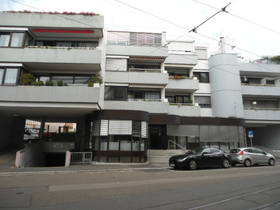 4-stöckige moderne Häuserzeile. In den Obergeschossen Balkone. Im EG links Einfahrt zur Tiefgarage. Rechts davon vorgelagerter Glas-Rundbau, daneben rechts 6 Stufen zu Glas-Schiebetüre.