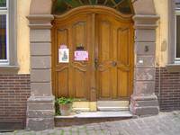 Auf dem Bild sieht man eine zweiflügelige Eingangstür, umgeben von einem steinernen Bogen und einer Stufe davor, die erkennbare Straße hat an dieser Stelle eine leichte Steigung, links an der Tür steht ein Blumentopf
