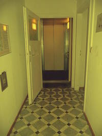 offene hölzerne Tür, dahinter die geöffneten Aufzugstüre und der Innenraum des Aufzuges, im Vordergrund ein Flur