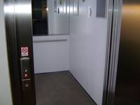 Edelstahl-Aufzugstür geöffnet. Links am Rahmen Tastaturuftaster,, Kabine mit dunklem Bodenbelag, Spiegel und Haltegriff an Rückwand, helle Wände.