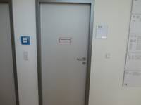 geschlossene hellgraue Tür mit einem dunklen Rahmen in einer weißen Wand. Links von der Tür ein blauer Feuermelder und eine weitere geschlossene Tür. Rechts neben der Tür hängen ein Türschild und Hinweisschilder mit der Raumverteilung 