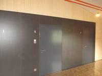 holzverkleidete Wand mit 3 Metalltüren, die Tür in der Mitte ist die Behindertentoilette. Neben der Tür ist ein Rollstuhlsymbol
