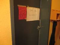 Dunkle Tür in einer gelben Wand. Auf die Tür sind 2 handschriftlich beschriebene Blätter geklebt: ein rotes mit verschiedenen Namen und ein größeres weißes mit der Aufschrift: Bitte leise hereinkommen