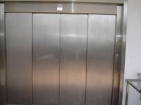 Im Bild ist die geschlossene metallische Eingangstür des Aufzuges zu sehen, die Tür besteht aus 4 Elementen und geht über die ganze Bildbreite, rechts ist der Taster zum rufen des Aufzuges erkennbar
