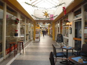 breiter Innenflur, beidseits Ladengeschäfte, im Vordergrund rechts blaue Stühle und Tisch eines Cafes. Glasdach über Passage.hts