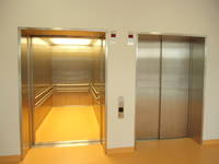 zwei Aufzüge, der linke ist offen der rechte ist geschlossen,  dazwischen Taste. Der offen stehende Aufzug ist beleuchtet; Kabien aus Metall mit Handlauf