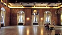 Großer Saal mit Holzparkettboden und großen Kronleuchtern. Große bogenförmige Fenster mit hellen Vorhangschals. Rechts im Saal steht ein Tisch mit weißer Tischdecke und sechs gepolsterten Stühlen