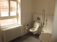 Eine weiße Hängetoilette an einer weiß gekachelten Wand. Haltegriffe auf beiden Seiten der Toilette. Links daneben ein großes Fenster, an der Wand rechts von Toilette hängt ein Waschbecken