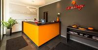 L-förmige orangefarbene Rezeption mit einer dunklen Deckplatte, davor zwei Teppeich, eine Topfpflanze und an der Wand ein kleines halbhohes Regal mit Broschüren, ausserdem an der Wand das Hotellogo