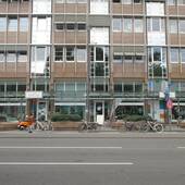 mehrstöckiges Gebäude, im Erdgeschoss Schaufensterfront, davor Pflanzenkübel, Fahrräder und die Straße