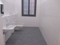 Länglicher Raum mit dunklem Bodenfliesen und weißen Wandkacheln. Auf der linken Seite ist ein Waschbecken, dahinter die Behindertentoilette mit weißen Haltegriffen rechts und links. In der schmalen hinteren Wand ist ein großes, außen vergittertes Fenster.