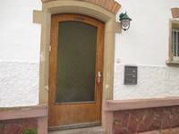 Tür mit großem, grünen Glaseinsatz in einem hellbraunen Holzrahmen. Die Fassade ist weiß mit einem Sandstein-Sockelber der Tür ist eine laternenförmige Lampe , rechts von der Tür sind Briefkästen