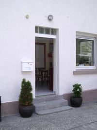 offene Tür in einem weißen Gebäude, vor dem Eingang zwei Stufen mit einem Blumentopf rechts und links, links an der Wand ist ein Briefkasten und rechts von der Tür ein Fenster, im Vordergrund ist ein Steinplatz